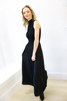 Woman wearing a black sleeveless dress standing against a white wall.