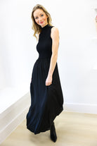 Woman wearing a black sleeveless dress standing against a white wall.