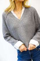 Person wearing a gray sweater with white cuffs and collar, blue jeans, and a necklace on a white background.