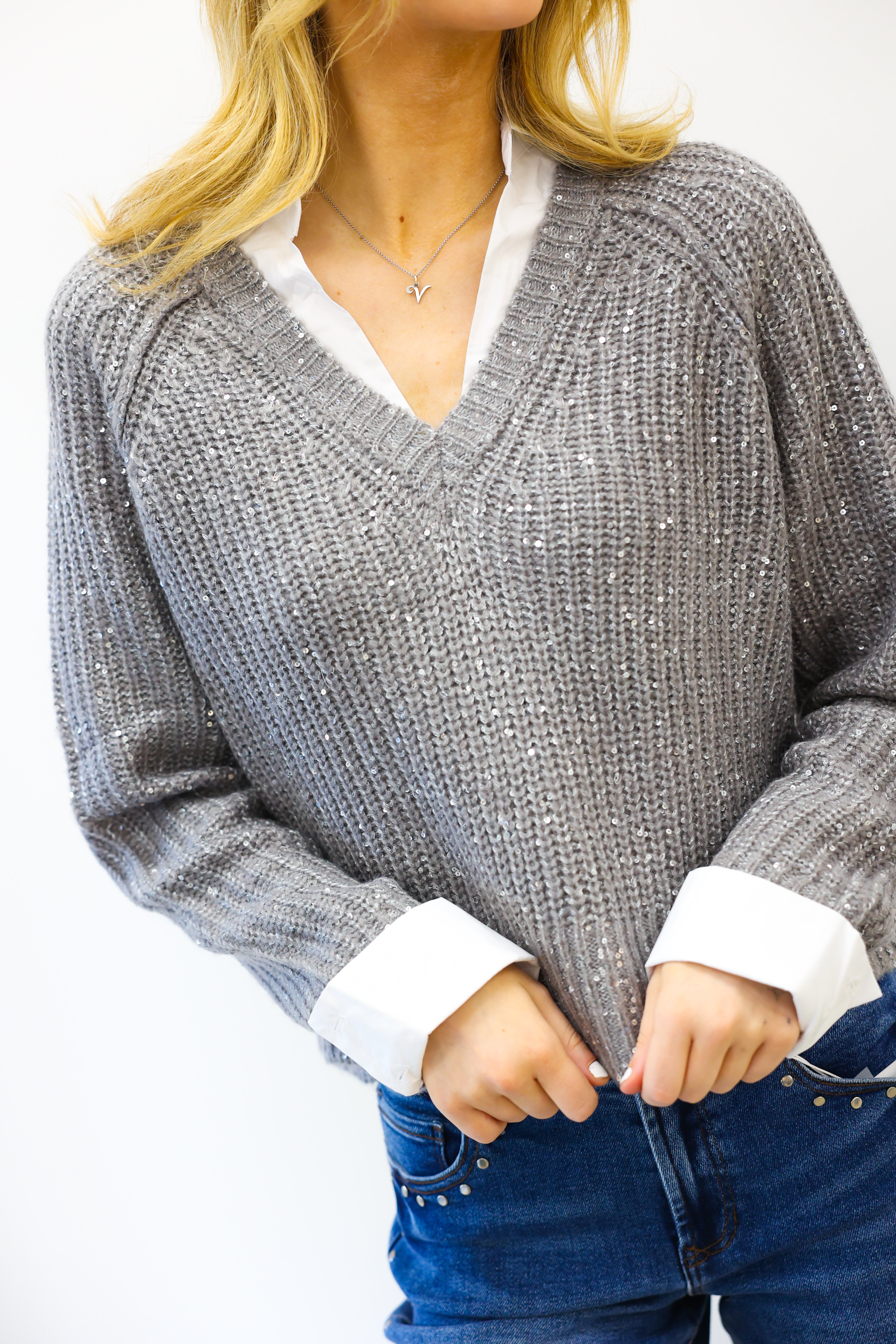 Person wearing a gray sweater with white cuffs and collar, blue jeans, and a necklace on a white background.