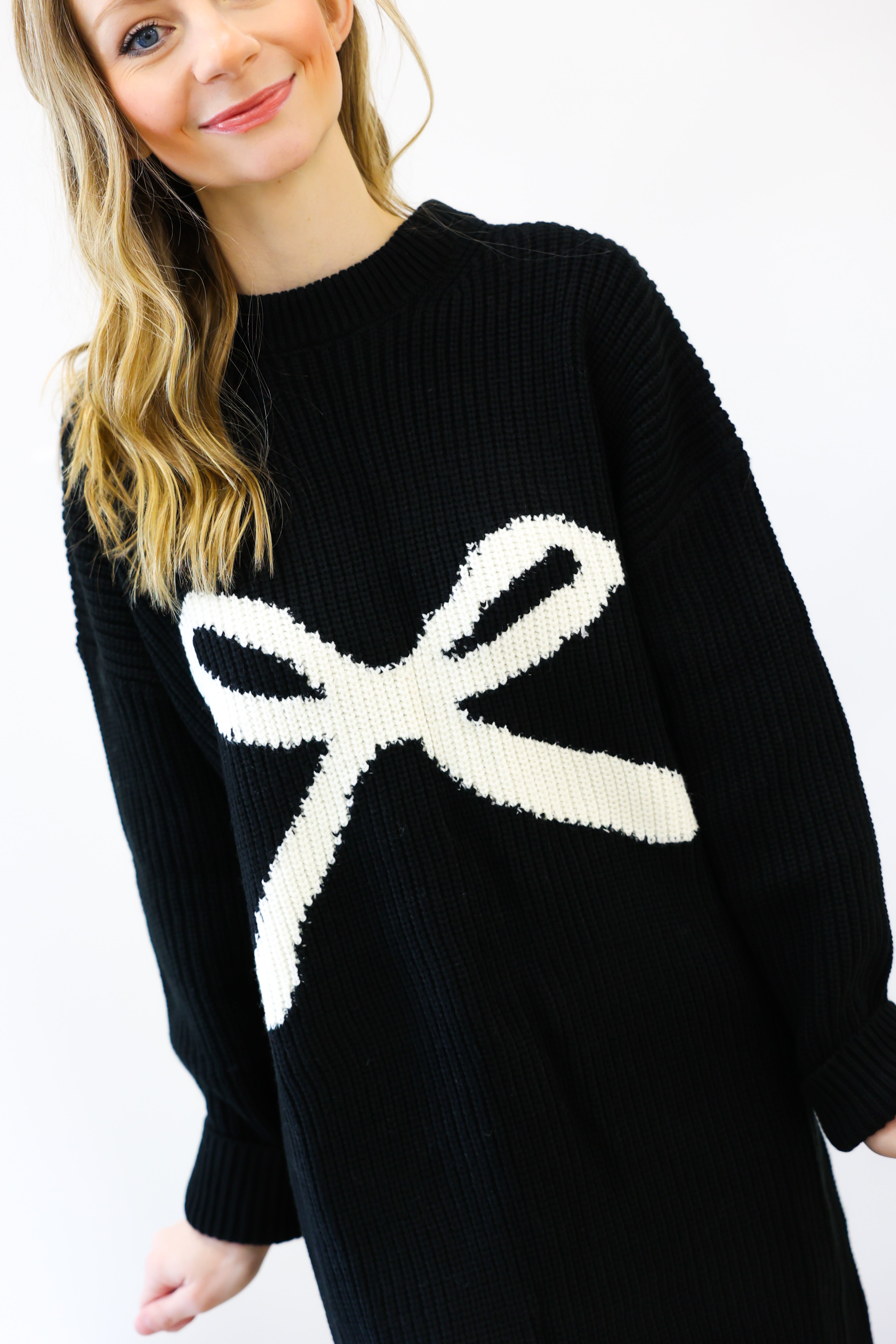 Black sweater with a large white bow design worn by a person on a white background