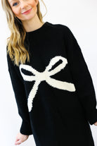 Black sweater with a large white bow design worn by a person on a white background