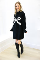 Woman wearing a black sweater dress with a white bow design and black knee-high boots on a white background.