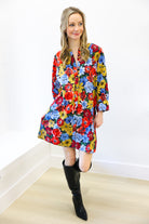 Woman wearing a colorful floral dress with black knee-high boots against a white wall.