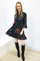 Woman wearing a dark floral dress with black knee-high boots against a white wall.