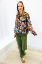 Woman wearing a colorful floral blouse and green pants against a white wall.