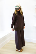 Person wearing a dark brown outfit with wide-leg pants and a cap indoors.