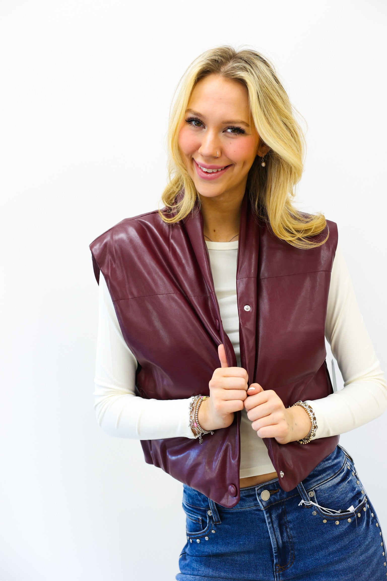 Woman wearing a burgundy leather vest over a white long-sleeve shirt and blue jeans on a white background
