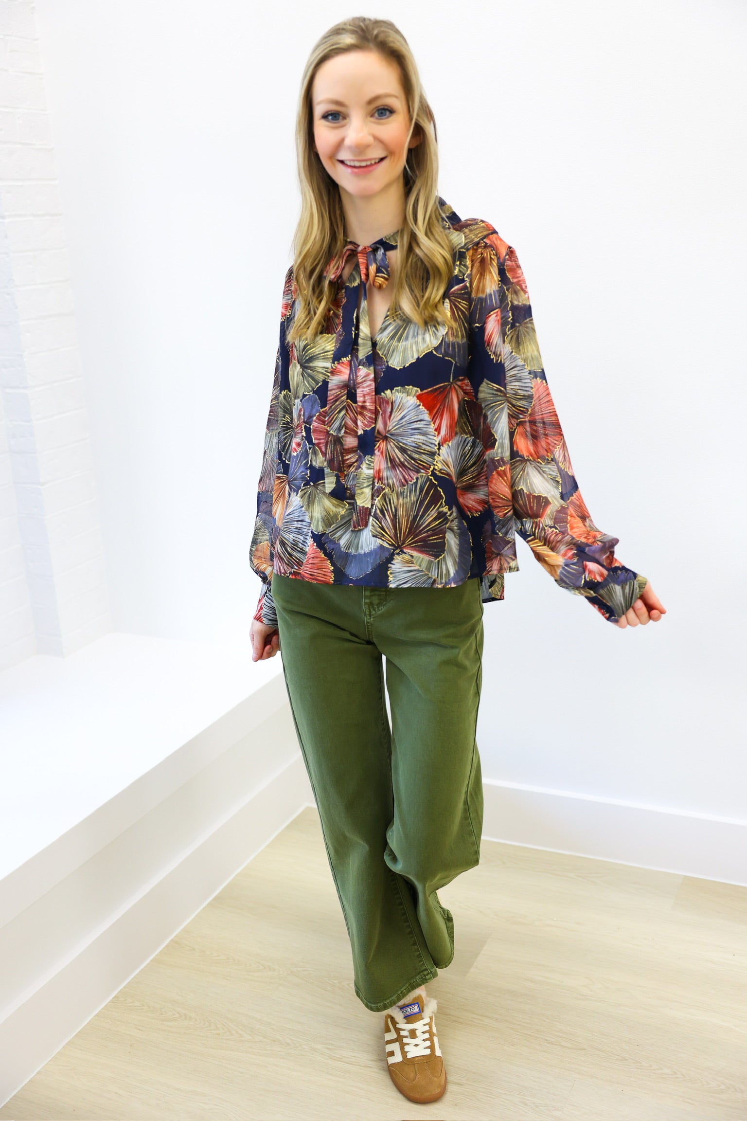Woman wearing a colorful floral blouse and green pants against a white wall.