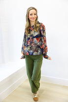 Woman wearing a colorful floral blouse and green pants against a white wall.