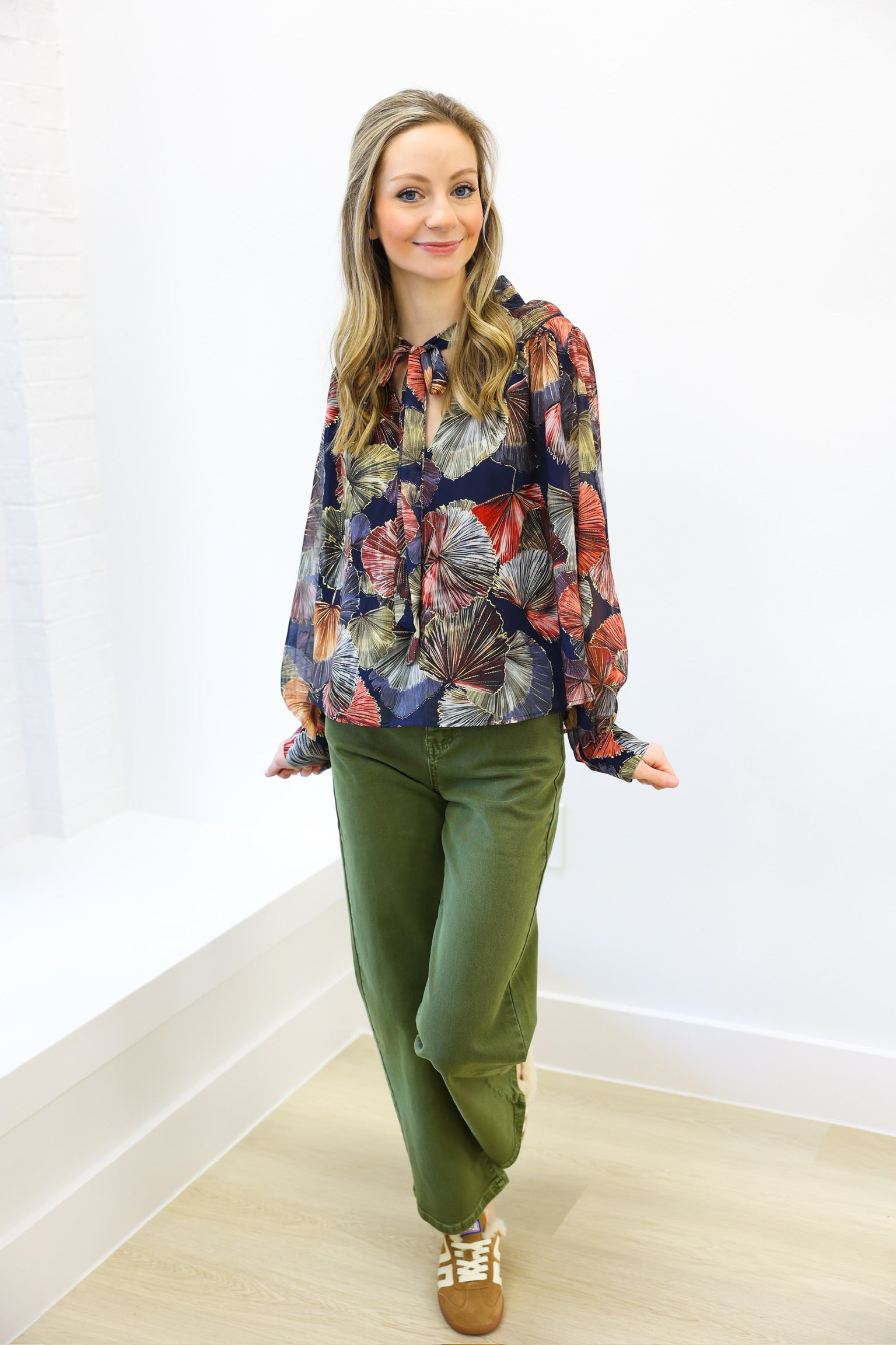 Woman wearing a colorful floral blouse and green pants against a white wall.