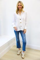 Woman wearing a white blouse with gold buttons, blue jeans, and white sneakers on a plain background