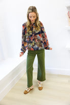 Woman wearing a colorful patterned top and green pants in a minimal indoor setting