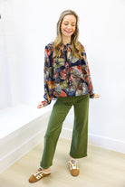 Woman wearing a colorful floral blouse and green pants against a white wall.