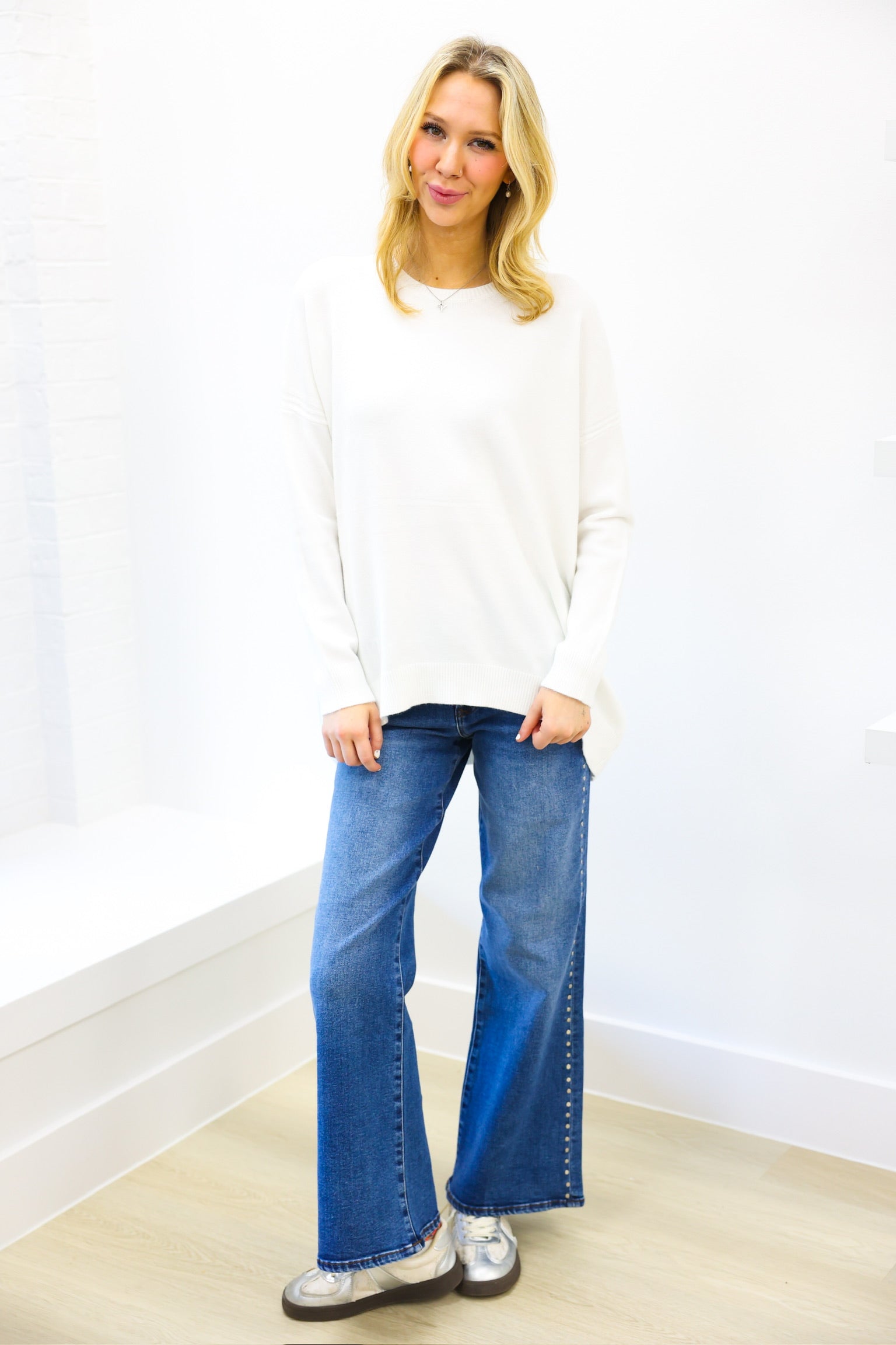 Woman wearing a white sweater and blue jeans standing against a white wall.