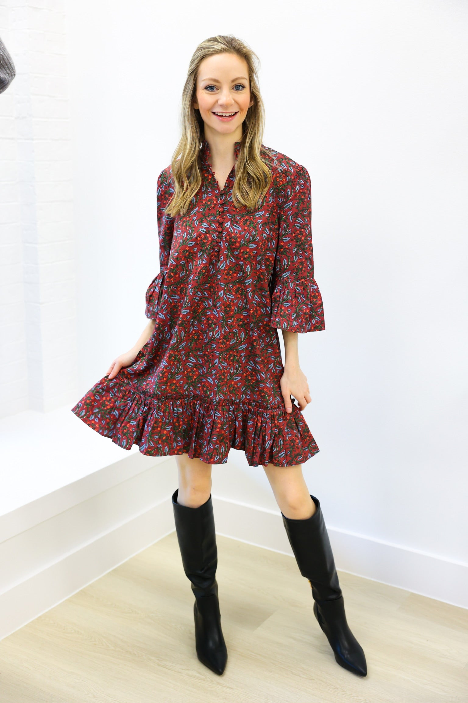 Woman wearing a patterned dress and black boots against a white wall.