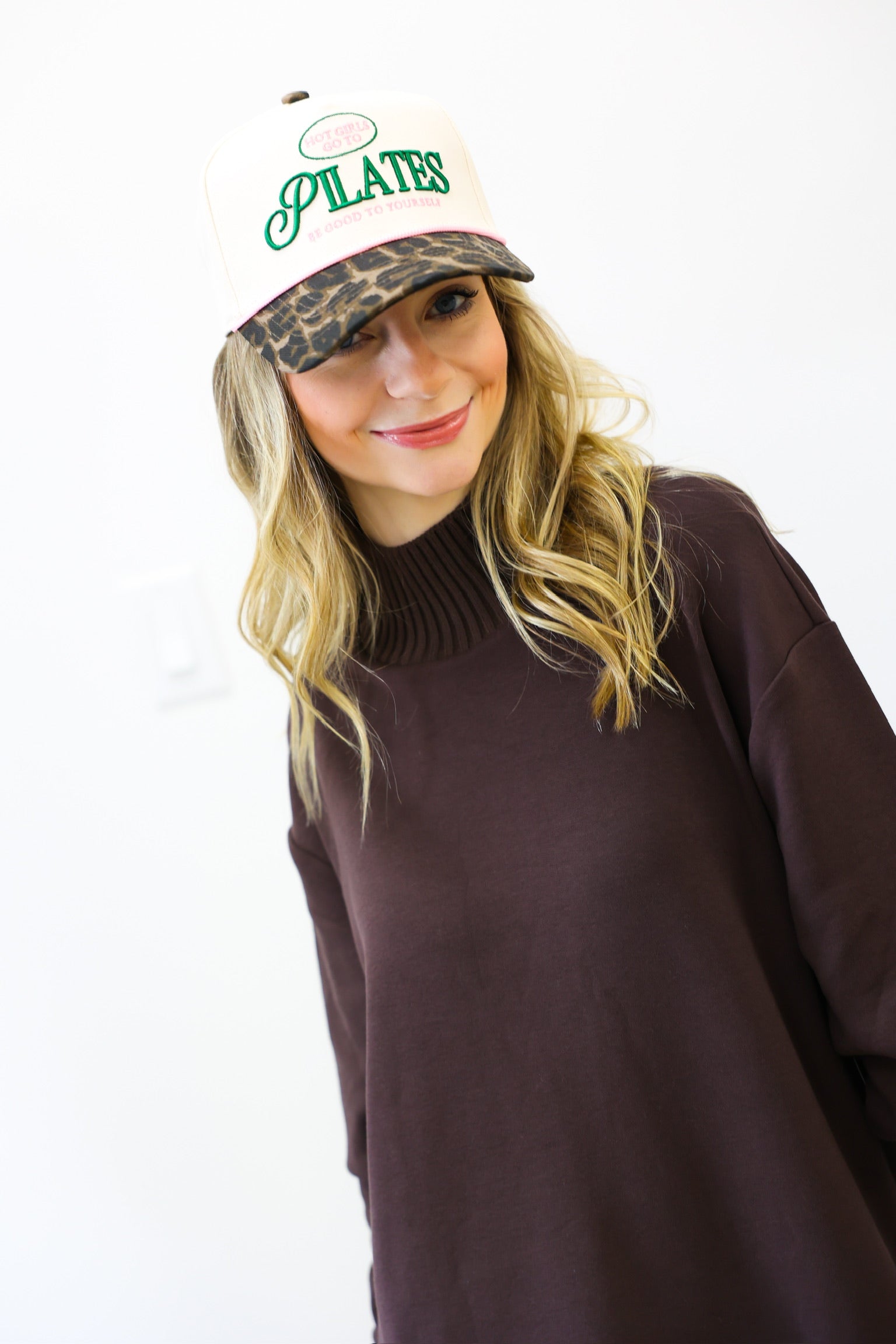 Woman wearing a brown sweater and a cap with 'Pilates' text on a white background