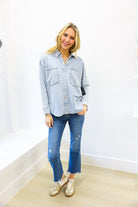 Woman wearing a light blue denim shirt and jeans standing against a white wall.