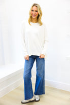 Woman wearing a white sweater and blue jeans standing against a white wall.