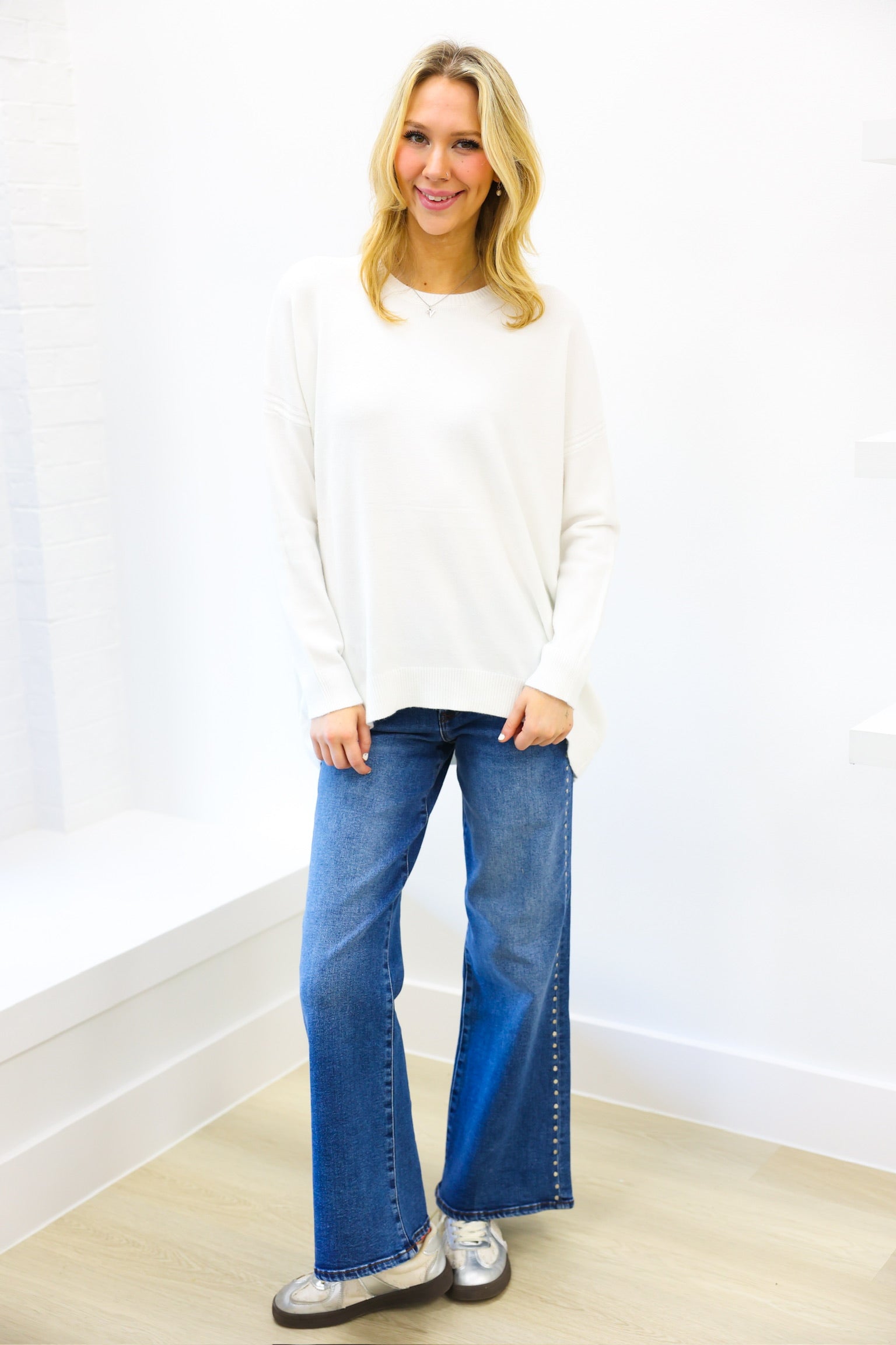 Woman wearing a white sweater and blue jeans standing against a white wall.