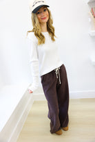Woman wearing a white long-sleeve top and brown pants against a white wall.