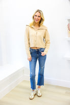 Woman wearing a beige jacket, blue jeans, and white sneakers in a minimalistic room.