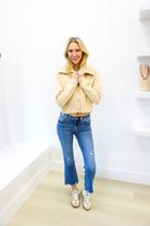 Woman wearing a beige jacket and blue jeans standing in a room with white walls and wooden floor.